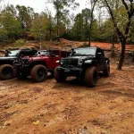 Three Jeeps with mud tires parked on a dirt trail surrounded by trees in a forested off-road park.
