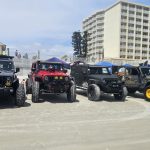 Row of modified Jeeps with tents behind them on the beach, with hotels and blue sky in the background.