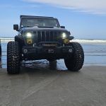 Black lifted Jeep Gladiator with headlights on, parked on wet sand by the ocean under a blue sky.