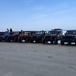 Group of Jeeps parked on the beach with members sitting on top, ocean waves in the background.