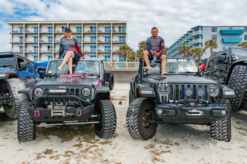Man and woman sitting on top of lifted Jeeps on the beach, with hotels and palm trees in the background.
